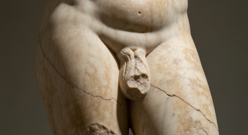 A detailed close-up of ancient marble statue genitalia with visible weathering and patina, showing artistic craftsmanship in natural museum lighting