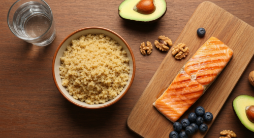A photorealistic overhead shot of a wooden table with a variety of heart-healthy foods: a bowl of quinoa, grilled salmon, avocado slices, a handful of walnuts, blueberries, and a glass of water. Natur