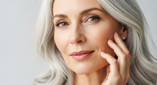 A close-up, photorealistic portrait of a woman around 55 years old, with elegant silver hair and healthy, glowing skin. She is gently touching her cheek, looking confident and natural. Soft natural li