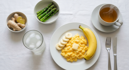 A photorealistic top-down view of a breakfast table with a plate of scrambled eggs, sliced banana, a glass of coconut water, a small bowl of steamed asparagus, and a cup of ginger tea. Soft morning li