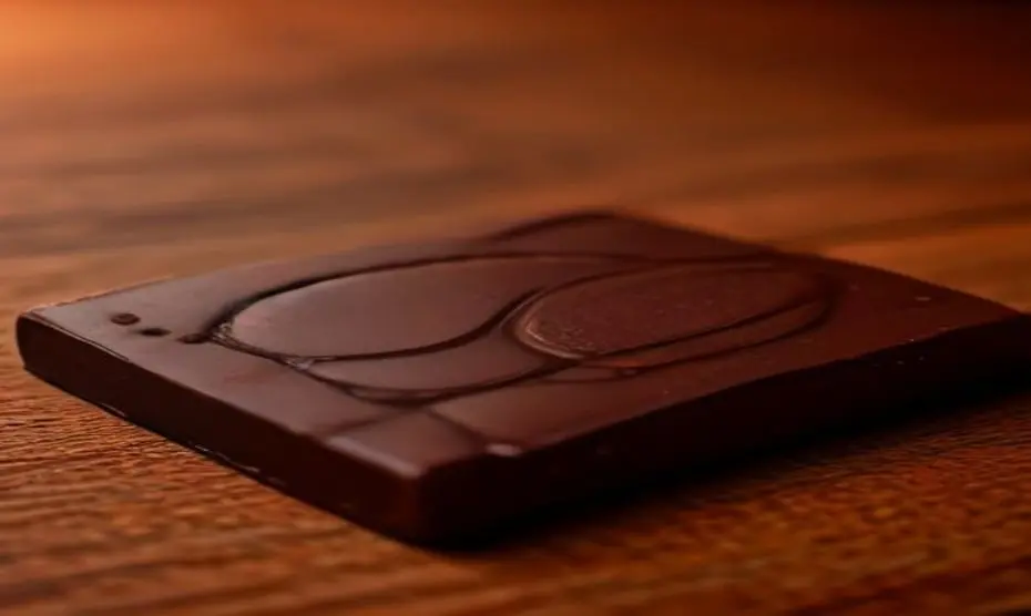 Cacao para la vista: alivia la fatiga digital 1 Photorealistic close-up of a dark chocolate bar with high cocoa percentage (85%) on a rustic wooden table, next to a pair of modern glasses reflecting a blue light screen, soft natural lighting, shall